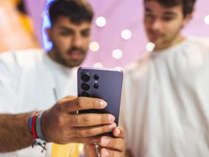 Close-up of the Samsung Galaxy S26 Ultra rear camera module in Cobalt Violet color, held by a young man while another looks on at a Samsung launch event with purple lighting in the background