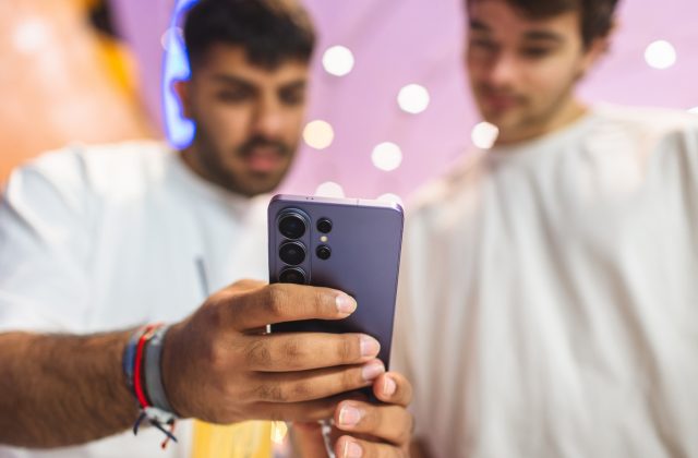 Close-up of the Samsung Galaxy S26 Ultra rear camera module in Cobalt Violet color, held by a young man while another looks on at a Samsung launch event with purple lighting in the background