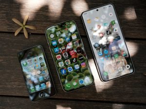 Three generations of smartphones on a wooden table — an older iPhone, a current-generation iPhone, and a Samsung Galaxy, illustrating how smartphone pricing and specs have evolved