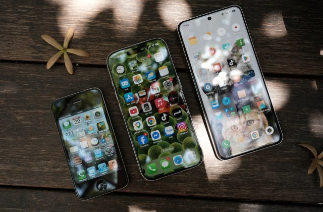 Three generations of smartphones on a wooden table — an older iPhone, a current-generation iPhone, and a Samsung Galaxy, illustrating how smartphone pricing and specs have evolved