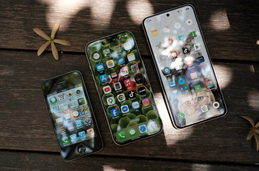 Three generations of smartphones on a wooden table — an older iPhone, a current-generation iPhone, and a Samsung Galaxy, illustrating how smartphone pricing and specs have evolved